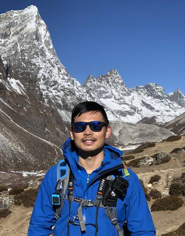 Man with sunglasses and blue jacket posing with snowy mountains in background.