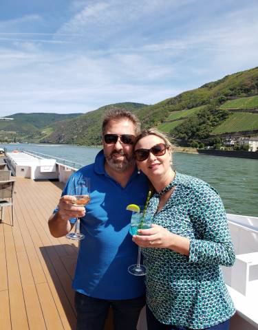 A couple enjoying drinks on a deck with a river and scenic hills.