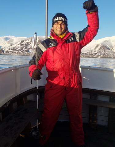 Person dressed in a red suit on a boat with snow-covered mountains in the background.
