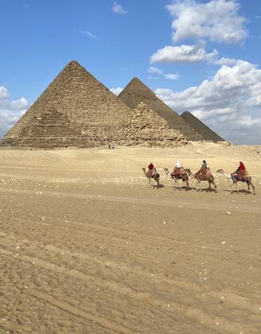 Camel riders in the desert with pyramids in the background.