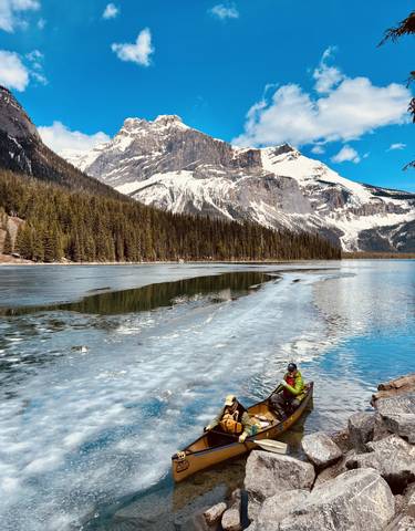 Person near a partially frozen lake with snow-capped mountains in the backdrop.