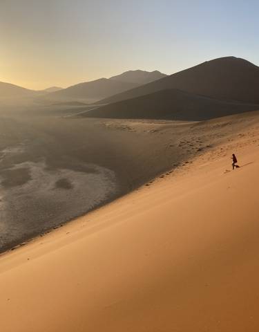 A person walking down a sand dune in the desert with the sun setting.
