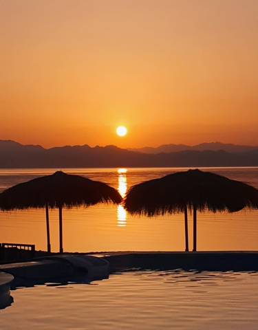 Sunset view over a calm ocean with silhouettes of distant mountains and beach umbrellas.