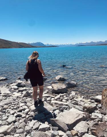 Person standing by the edge of a clear blue lake with mountains in the background.