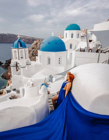 Woman in a blue dress sitting by white buildings with blue domes overlooking the sea.