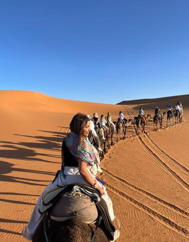 Line of camels and riders in a desert setting with sand dunes.