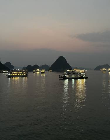 Boats with lights on a tranquil bay during twilight, likely in Halong Bay, Vietnam.