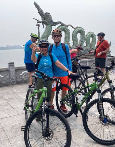 Group of cyclists posing in front of a scenic lake with a dragon sculpture.
