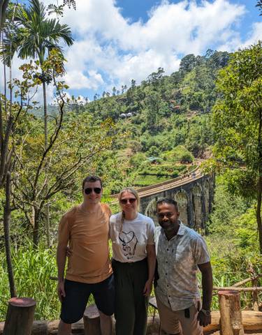 Three people posing in front of a railway bridge and lush landscape.