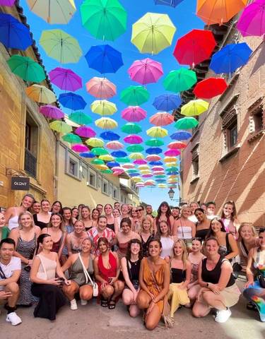 Large group of people posing under colorful umbrellas in a narrow street.
