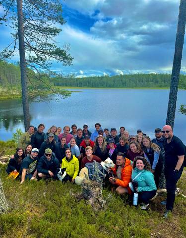 Large group of people posing by a serene lake in a forest.