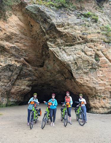 Four bikers in front of a sandstone cave with carvings.