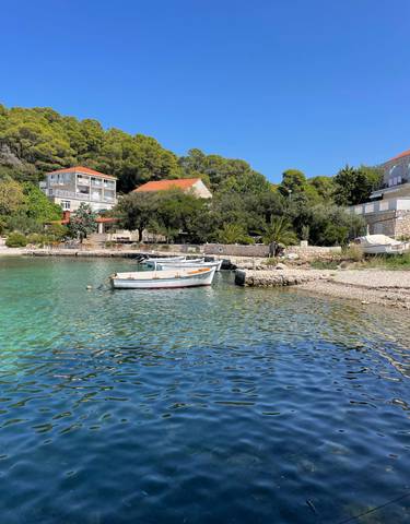 Small boats floating on clear turquoise water near a pebble beach with houses and trees in the background.