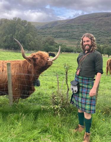 Man in traditional Scottish attire with a Highland cow in the background.