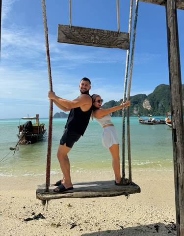 Two people on a swing by the beach with boats in the background.