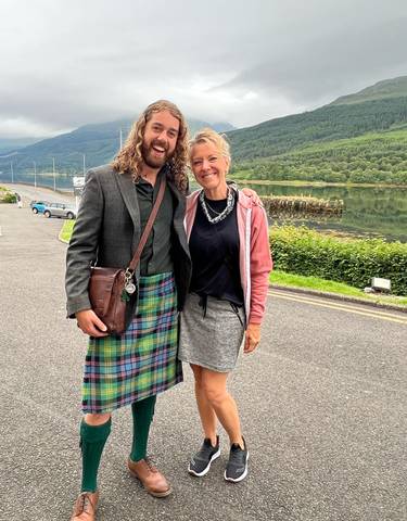 Two people smiling, one wearing a kilt, with a scenic winter countryside backdrop.