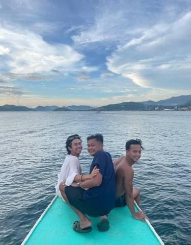 Three people smiling in front of a scenic water body with distant hills under a clear sky.