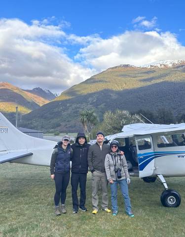 Four people standing in front of a small airplane with mountain scenery in the background.