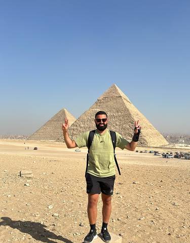 Person posing with pyramids in the background on a clear day.