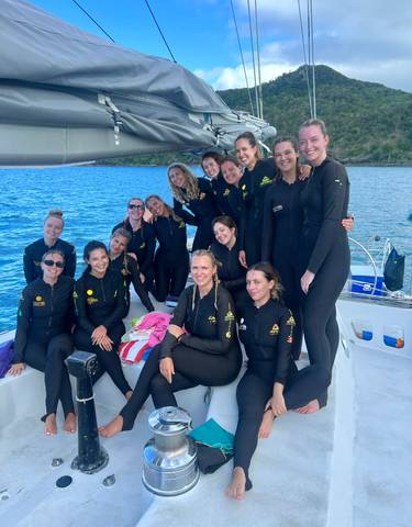 A group dressed in wetsuits posing on a sailboat on the ocean.