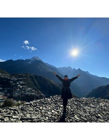 A person standing with arms raised on a hill with mountain vistas.