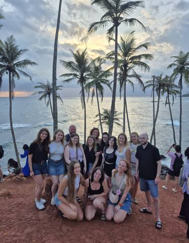 Group of people posing with palm trees and ocean at sunset.