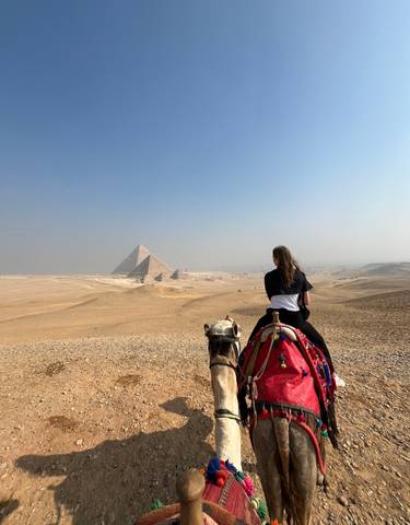 Person on a camel looking at the pyramids in a desert landscape.