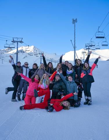 A group of people in colorful winter clothing celebrating on a snowy slope with mountains and ski lifts.
