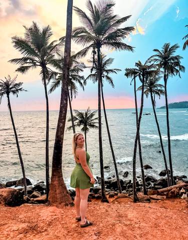 Woman posing by the beach with palm trees and a colorful sunset.