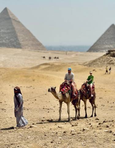 Tourists riding camels in the desert with pyramids in the background.
