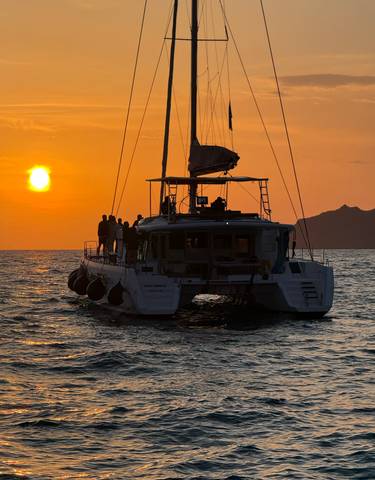 Silhouetted catamaran at sunset with people on board.