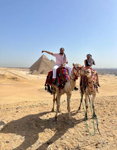 Two people on camels with pyramids in the background.