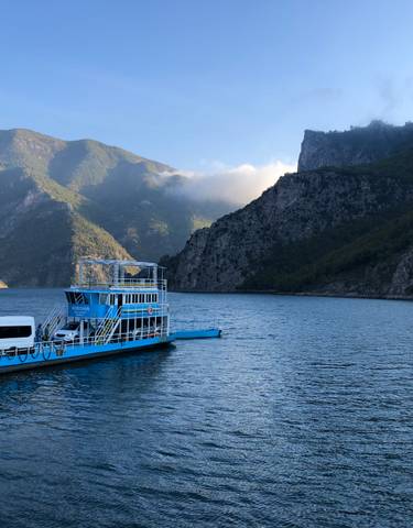 Blue ferry carrying a van on a scenic lake surrounded by mountains.