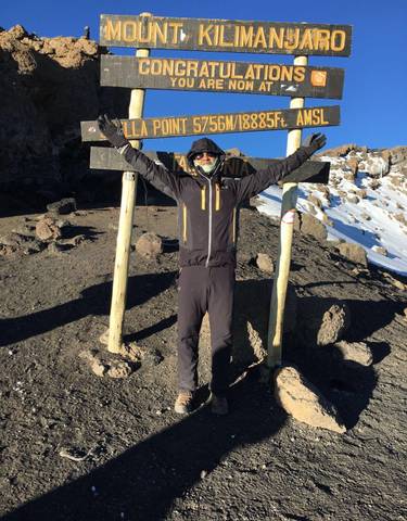 A person posing triumphantly on a mountain summit with a sign.