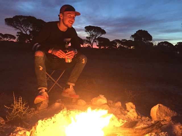       Person sitting by a campfire at sunset with trees in the background.
  