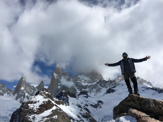       Person standing with open arms against a backdrop of snow-capped mountains.
  