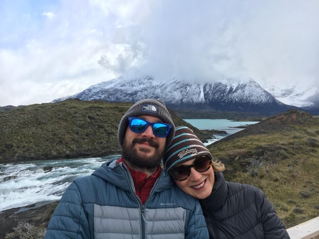       Two people smiling outdoors with scenic mountains and a river.
  