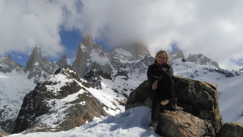       Person sitting on a rock with a snowy mountain backdrop partially covered by clouds.
  