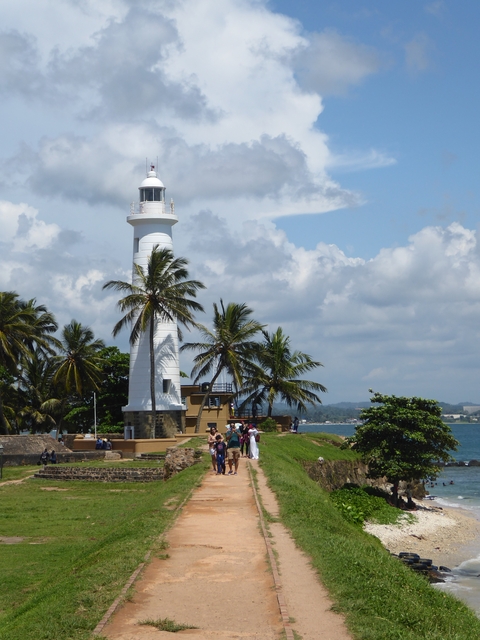 White lighthouse with palm trees and people nearby.