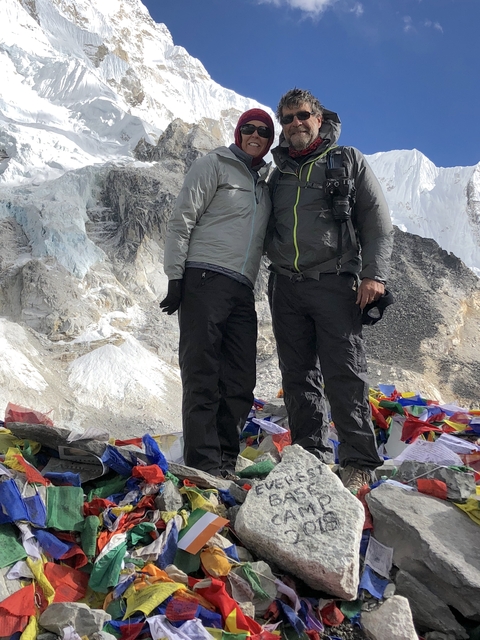 Two people standing amidst colorful prayer flags and mountains.