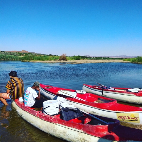 People sitting with kayaks on the bank of a river.