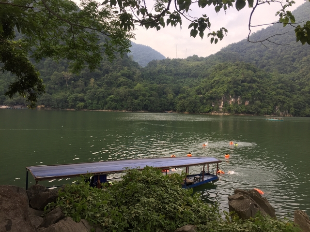 Boats with people swimming in a lake surrounded by green mountains.