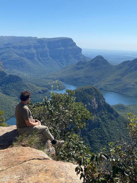 A person sitting on a cliff with a panoramic view of a valley and mountains.