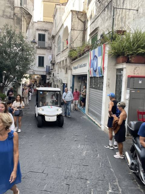       Cobbled street scene with people and a small vehicle.
  
