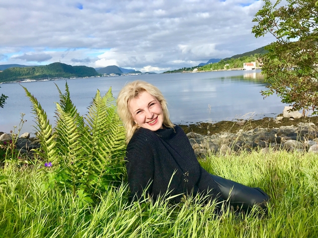 A woman sitting in grass with mountains and water in the background.