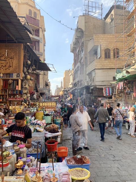 Busy market street with vendors and shoppers under a blue sky.