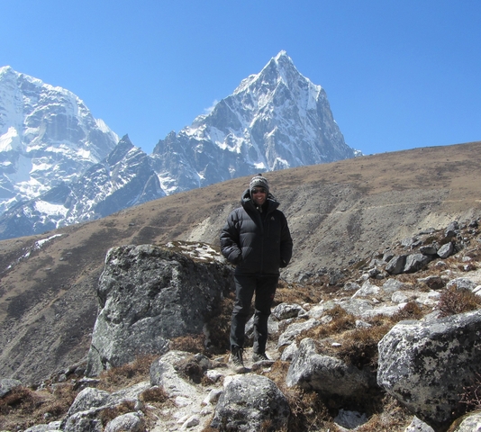       A person standing on a rocky landscape with snow peaks.
  