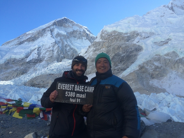 Two people holding a sign at Everest Base Camp with snowy mountains.