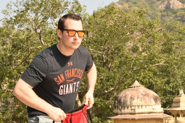 A person wearing sunglasses and a San Francisco Giants shirt with trees and domed architecture in the background.