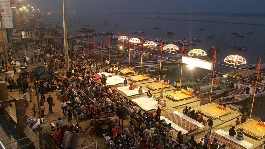 Crowded riverside ceremony during the evening with boats in the background.